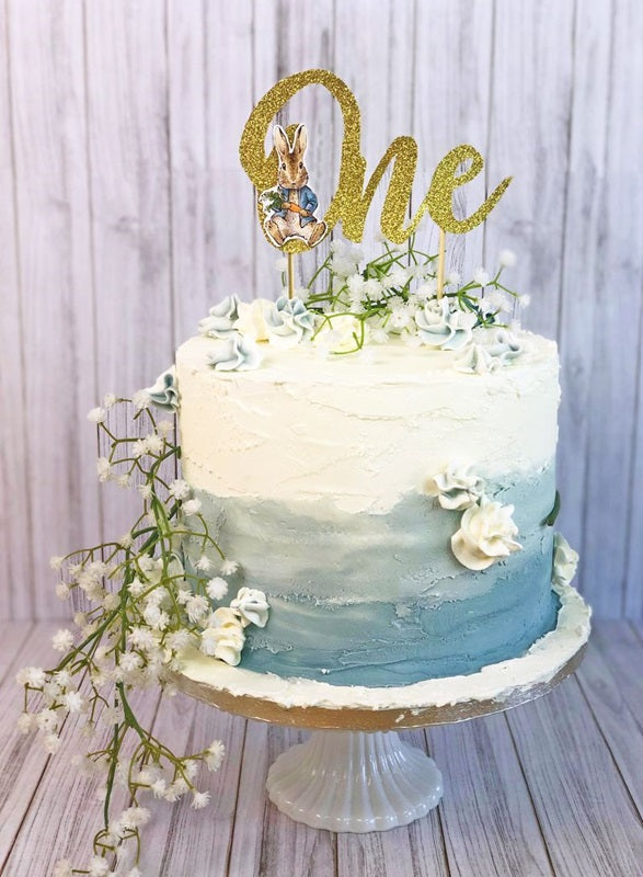 Example of Use - The Peter Rabbit Gold Glitter "One" cake topper atop a White with Blue Ombre buttercream cake adorned with White and green flowers and foliage.